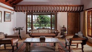 Quarto tradicional com teto de madeira, uma janela ampla com vista para o verde, um sofá central e duas mesas de apoio.