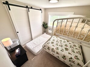 Ein Schlafzimmer mit einem Einzelbett, einem kleinen Nachttisch, einem Fenster mit Rollläden und einer Tür mit schwarzem Griff.