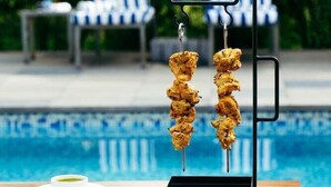 A poolside setting with a grill stand holding skewers of food, a table with a plate and a cup, and a lounge chair in the background. - De Garland, Palolem - AM Hotel Kollection (Canacona)