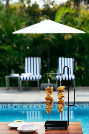 A poolside setting with a grill stand holding skewers of food, a table with a plate and a cup, and a lounge chair in the background. - De Garland, Palolem - AM Hotel Kollection (Canacona)