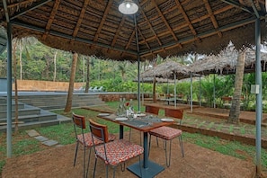 An outdoor dining area with a thatched roof, wooden tables, and chairs. - De Garland, Palolem - AM Hotel Kollection (Canacona)