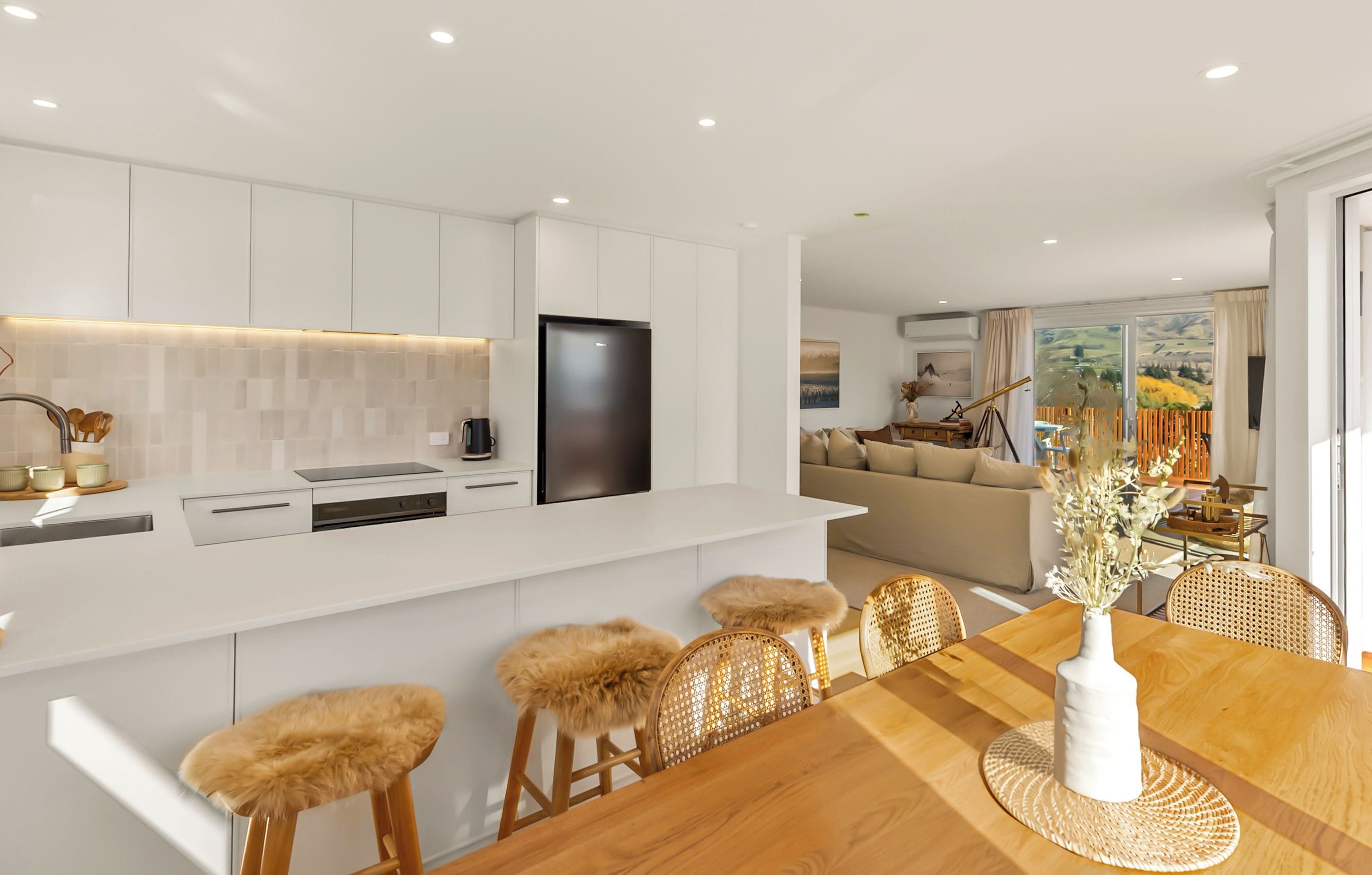 A modern kitchen with white cabinetry, a black refrigerator, and a dining area with a wooden table and rattan chairs.
