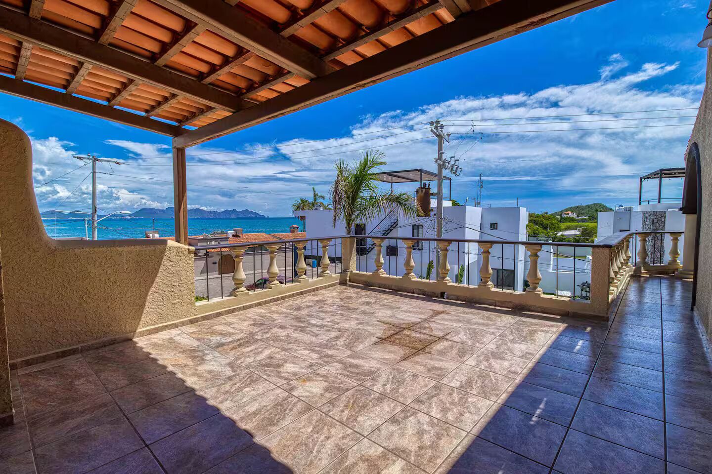 A covered outdoor terrace with a view of the sea and mountains, featuring a tiled floor and a railing.