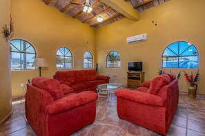 A spacious living room with a red sofa set, a glass coffee table, and a TV on a wooden cabinet. There are arched windows providing natural light and a view outside. - Ocean views, close to beach and marina boardwalk! (San Carlos Nuevo Guaymas)