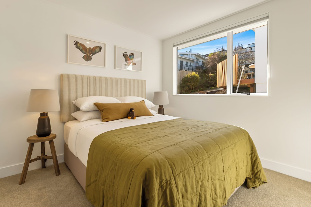 Un dormitorio con cama, almohadas, lámpara, ventana con vistas y dos cuadros en la pared.