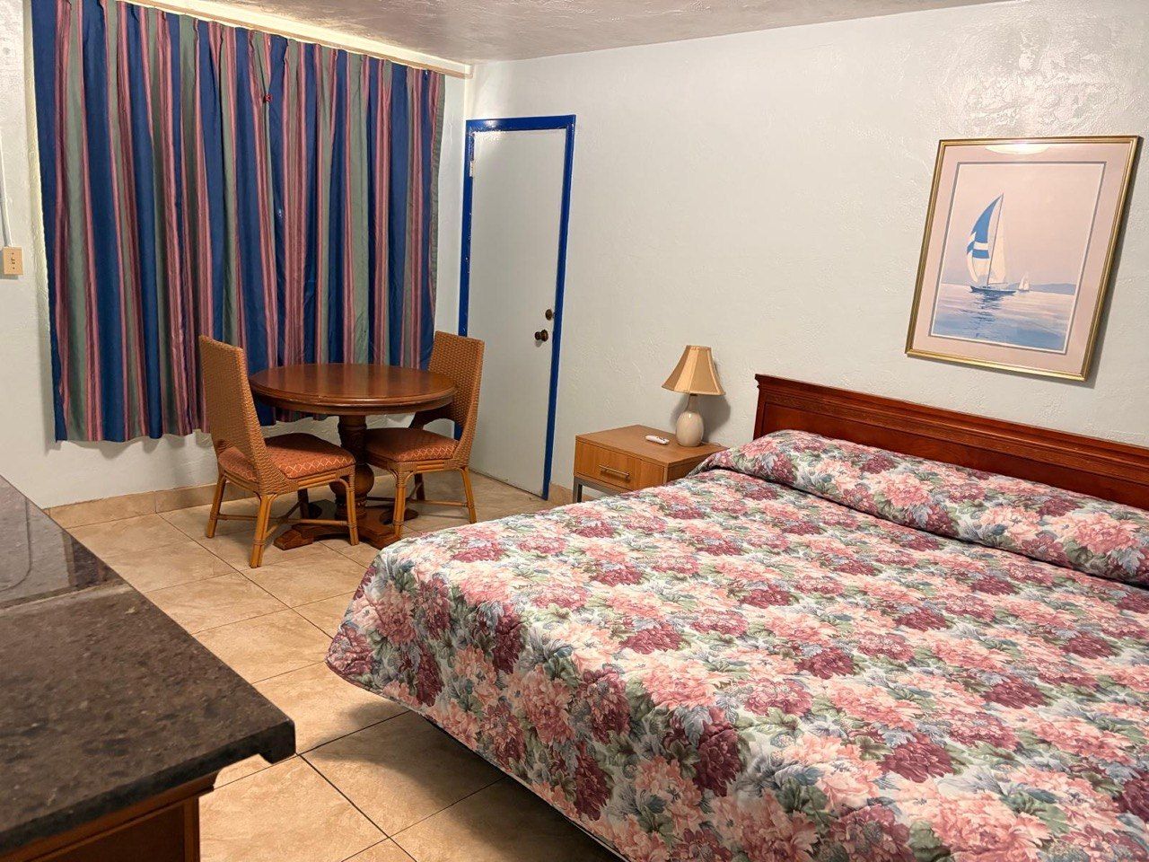 A bedroom with a bed, a wooden table with chairs, a nightstand, and a door.