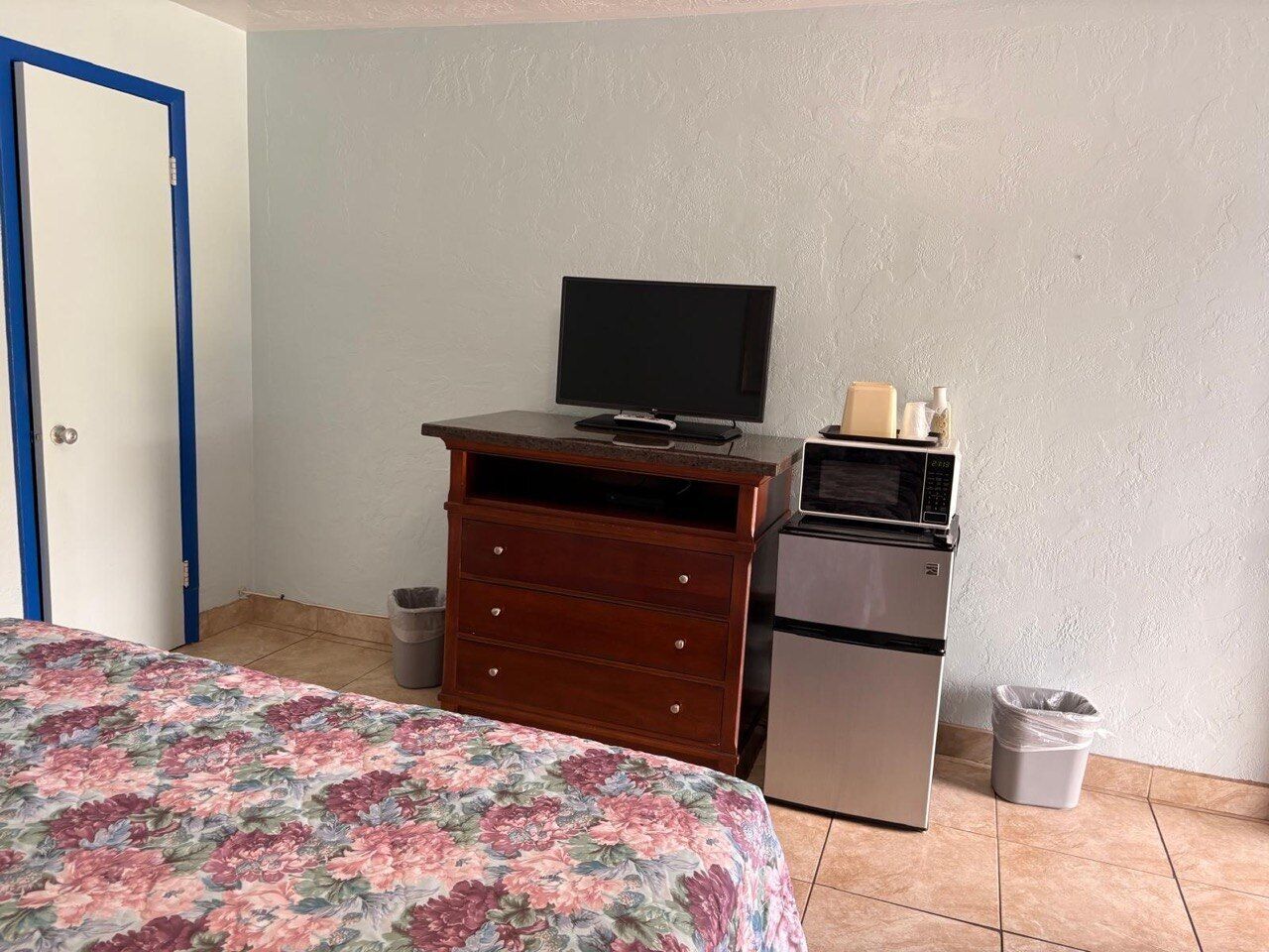 A bedroom with a bed, a dresser, a television, a microwave, and a refrigerator.
