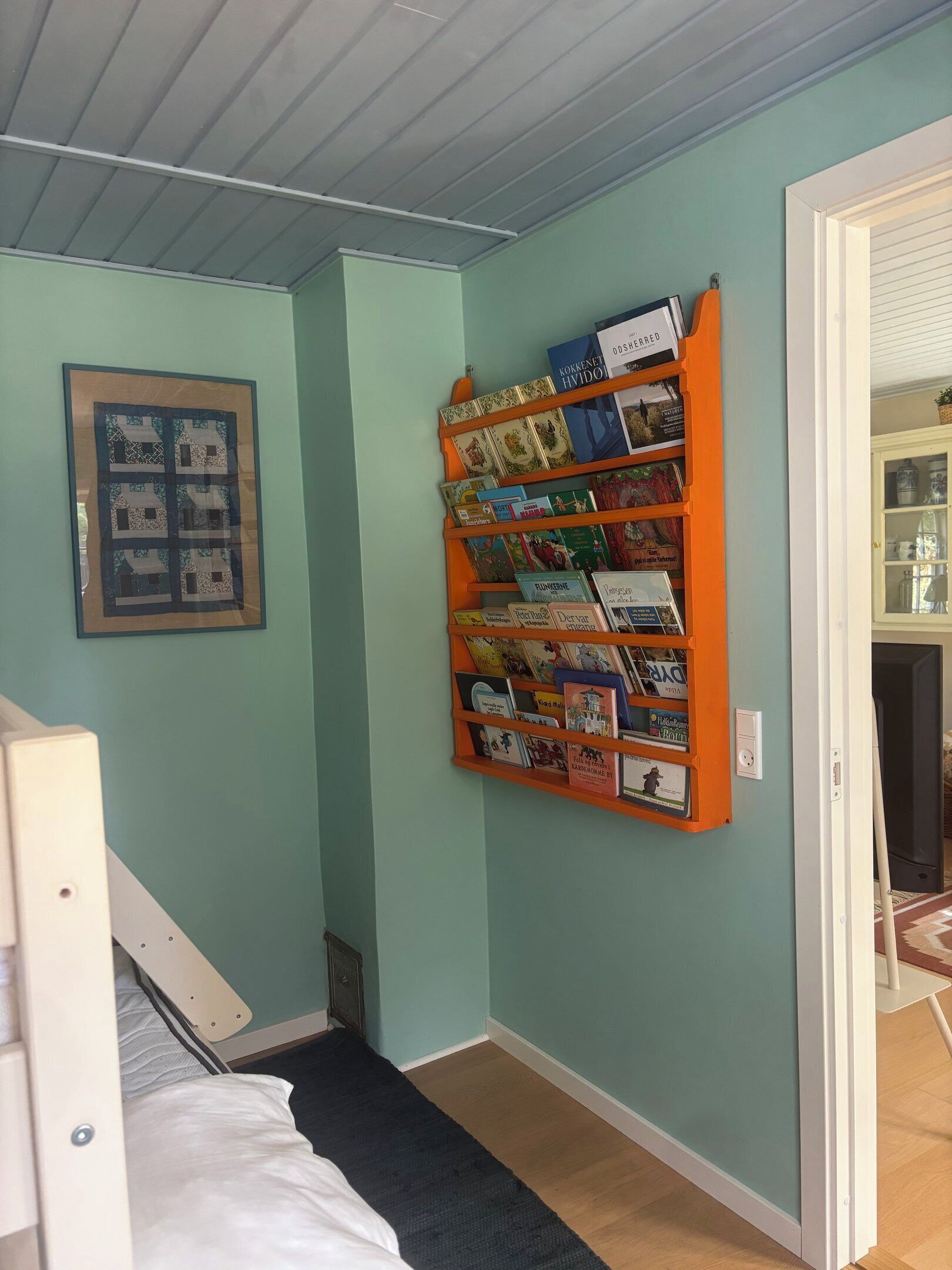 Una habitación con una estantería llena de libros, un cuadro en la pared y una cama con edredón blanco.