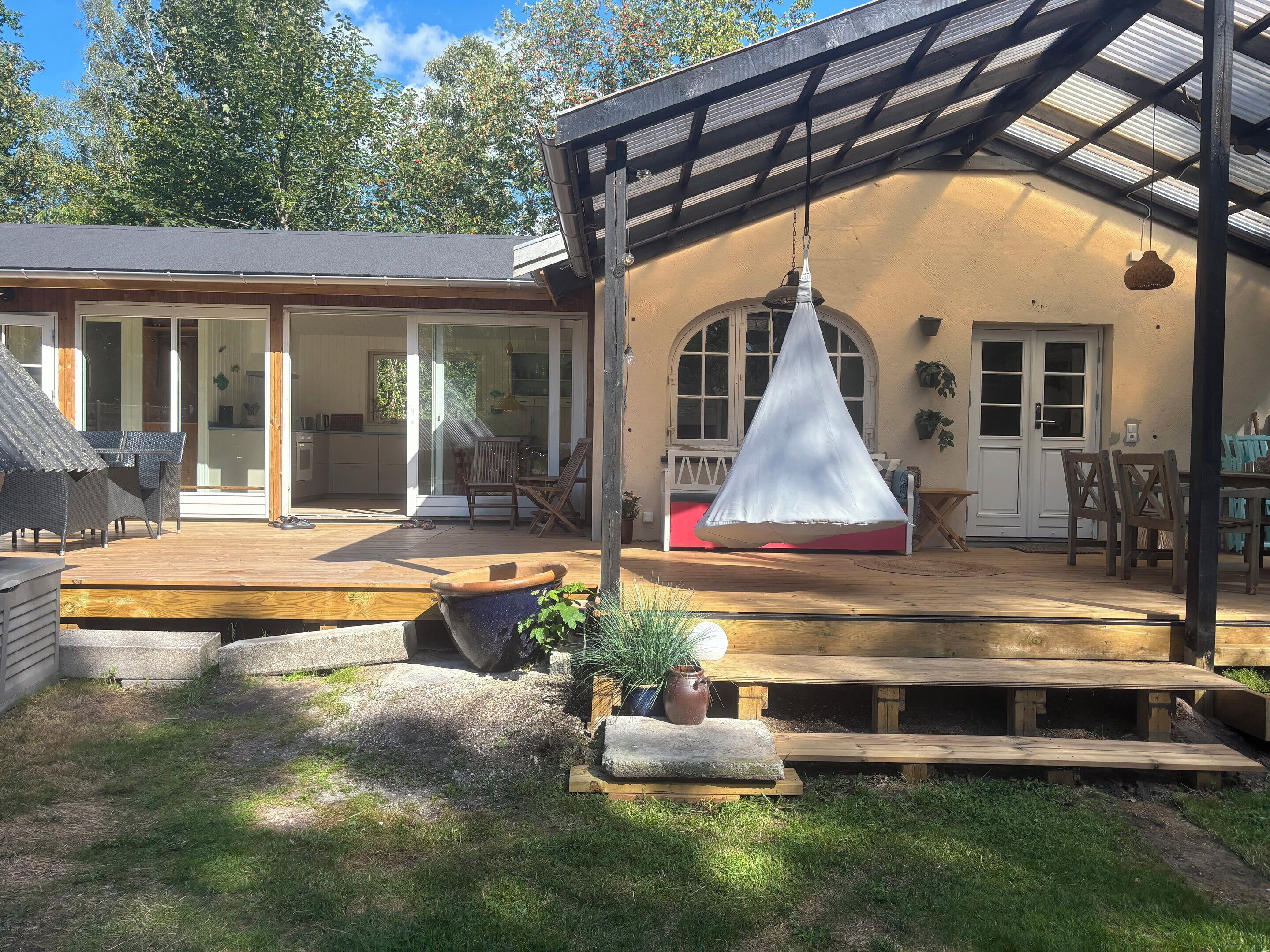 Un espacio exterior techado con cubierta de madera, una lámpara colgante y vista a una casa con ventanas amplias y una puerta.