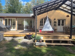 Ein überdachter Außenbereich mit Holzterrasse, einer hängenden Leuchte und Blick auf ein Haus mit großen Fenstern und einer Tür.