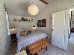 A bedroom with a bed, bedside tables, a bench, a hanging light fixture, a door, and a shelf with decorative items. - Lovely summer house from 2025 in Rørvig (Nykøbing Sj)
