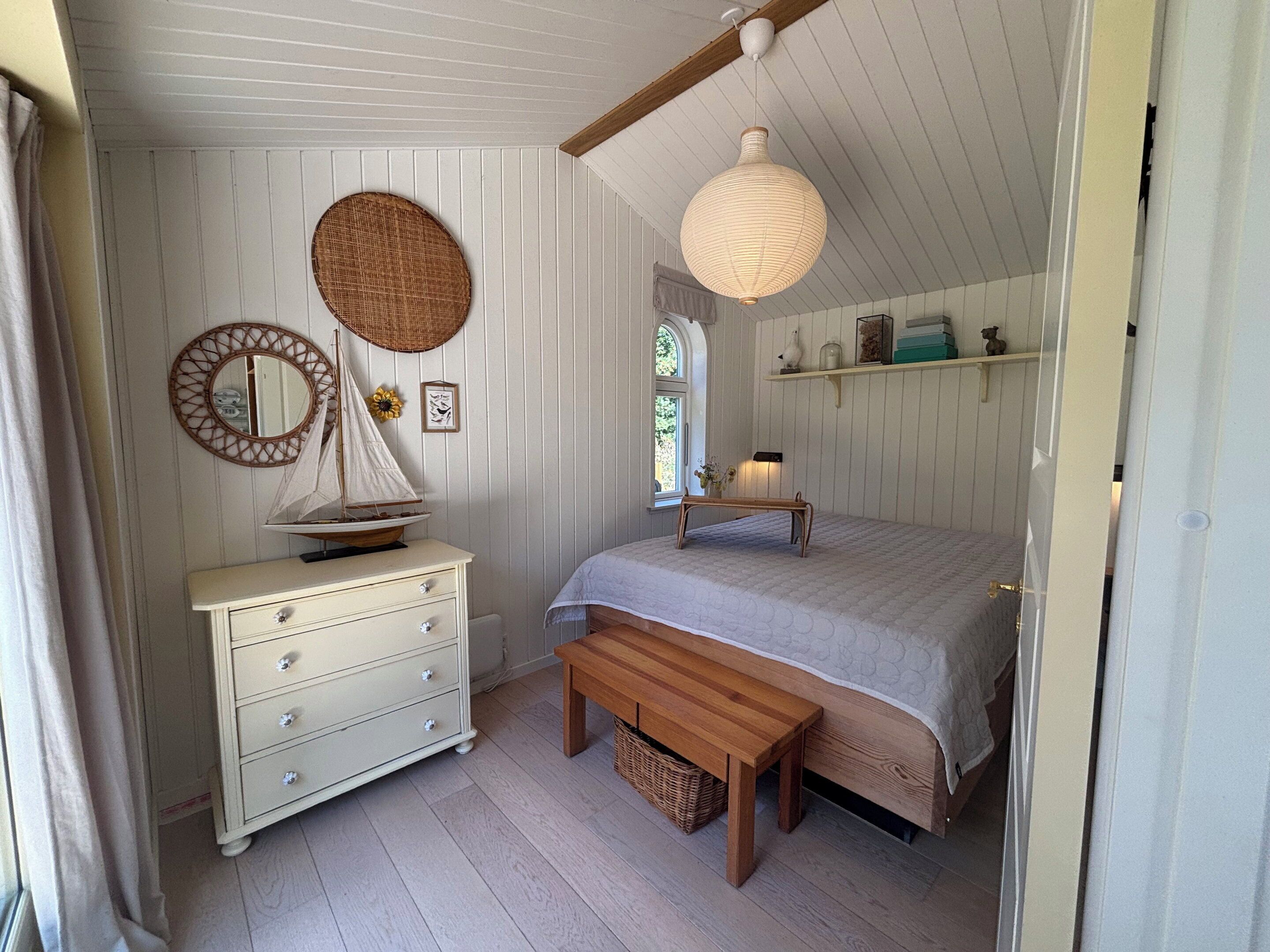 A bedroom with a bed, a dresser, a bench, a hanging light fixture, and a wall-mounted mirror.