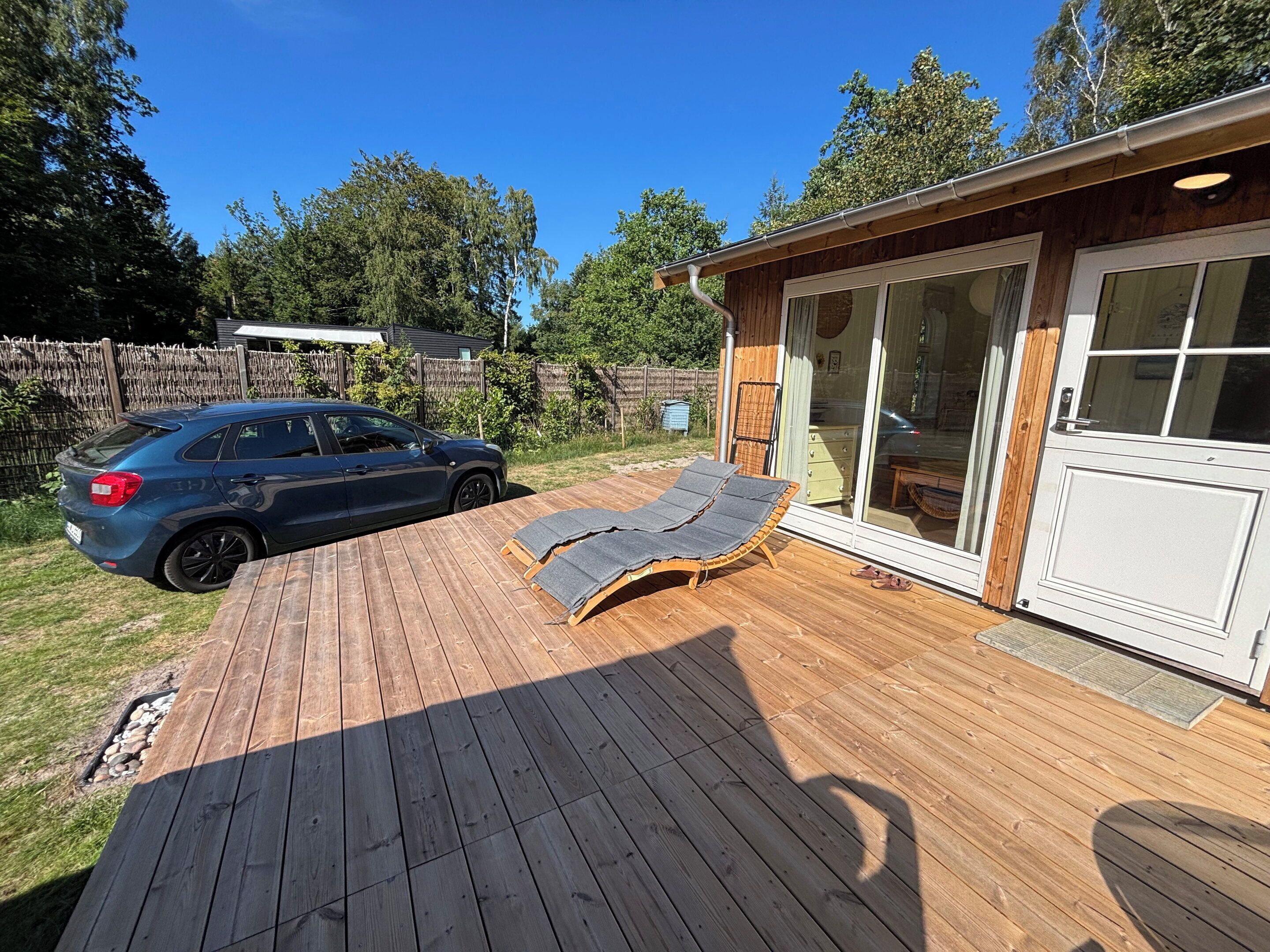 Une terrasse en bois avec des chaises longues, une voiture et un bâtiment doté d’une porte-fenêtre coulissante.