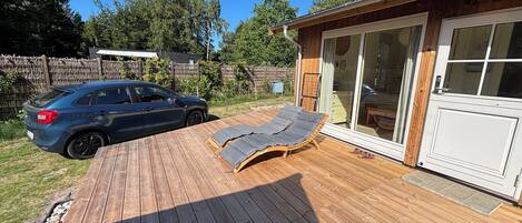 Une terrasse en bois avec des chaises longues, une voiture et un bâtiment doté d’une porte-fenêtre coulissante.
