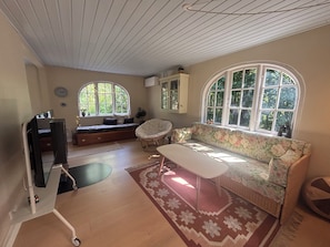 A room with a sofa, a chair, a table, and a window with a view of greenery. - Lovely summer house from 2025 in Rørvig (Nykøbing Sj)