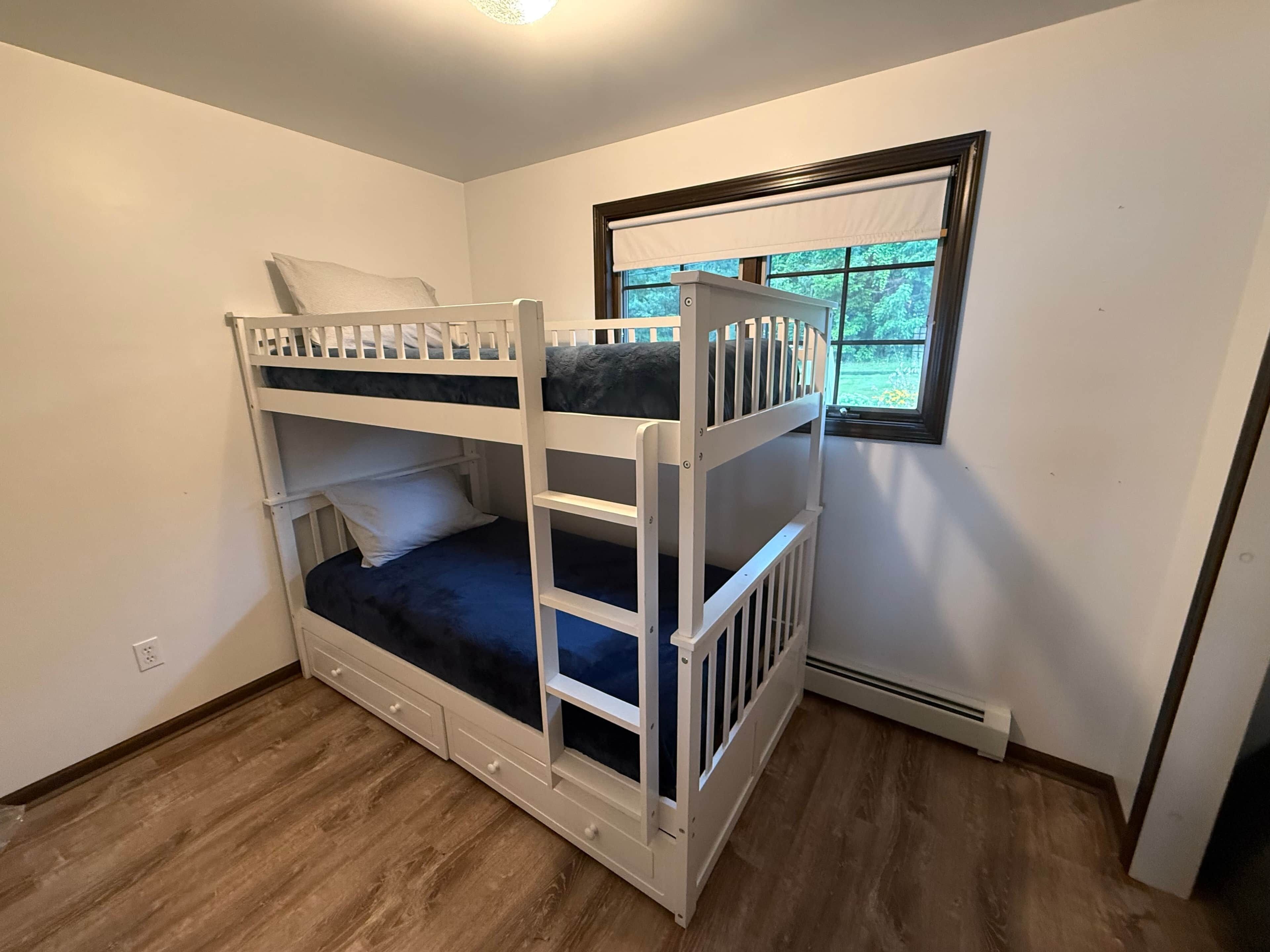 Une chambre avec un lit superposé, une fenêtre, un plancher en bois et un mur blanc.