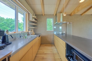 A modern kitchen with wooden cabinets, a stainless steel countertop, and a window providing a view of greenery outside. - Ume House (Kutchan)