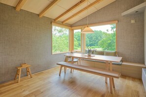 A room with a wooden table, benches, and a view of greenery through large windows. - Ume House (Kutchan)