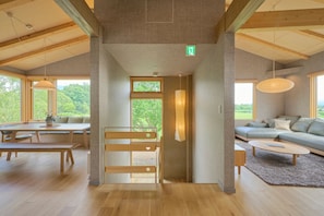 A spacious room with a wooden floor, a sofa, a table, and a bench. There is a large window providing a view of greenery outside. - Ume House (Kutchan)