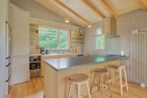 A modern kitchen with a central island, bar stools, wooden beams, and a view of greenery outside. - Ume House (Kutchan)