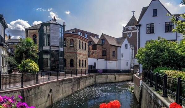 A canal with buildings on either side, a bridge, and a small boat with red flowers. - Beautiful Riverside Home Close to Kingston Town pet friendly (Kingston upon Thames)