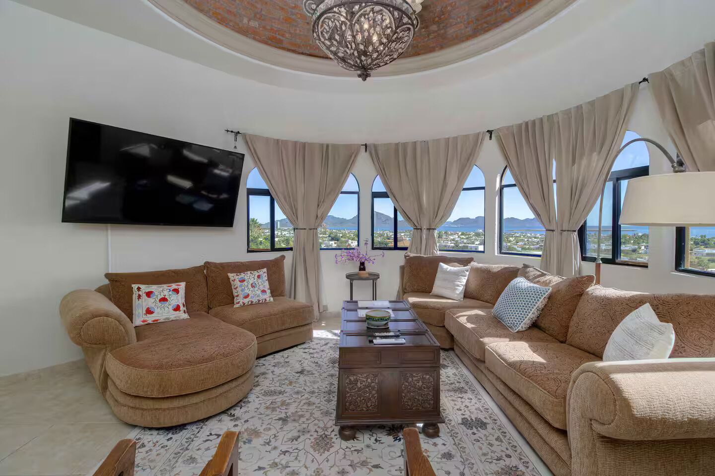 A spacious living room with a large flat-screen TV, comfortable sofas, a coffee table, and a view of the landscape through large windows.
