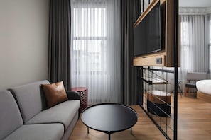 A modern living room with a sofa, a round coffee table, a flat-screen TV mounted on a wooden panel, and a bed in the background. - Moxy Istanbul Beyoglu (Istanbul)