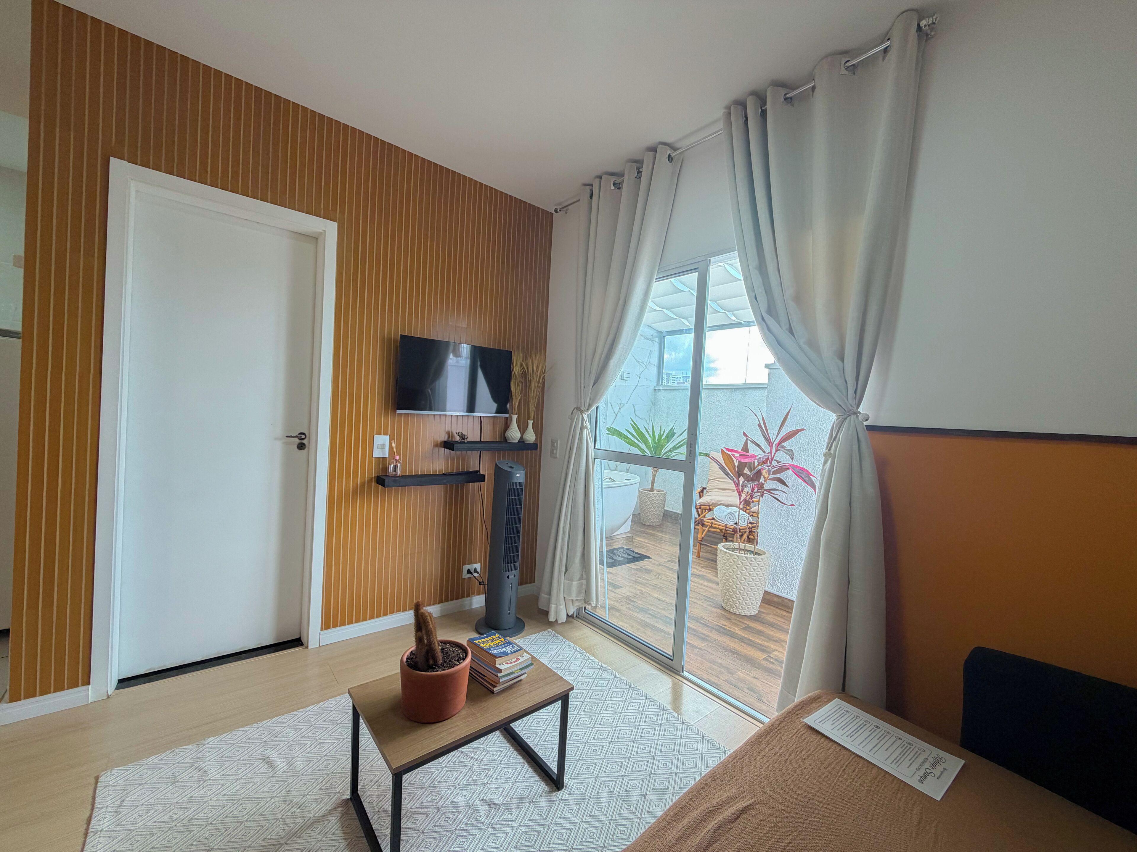Quarto de hotel moderno com televisão, uma pequena mesa com livros, uma planta em vaso e vista para o exterior através de uma porta de vidro deslizante.