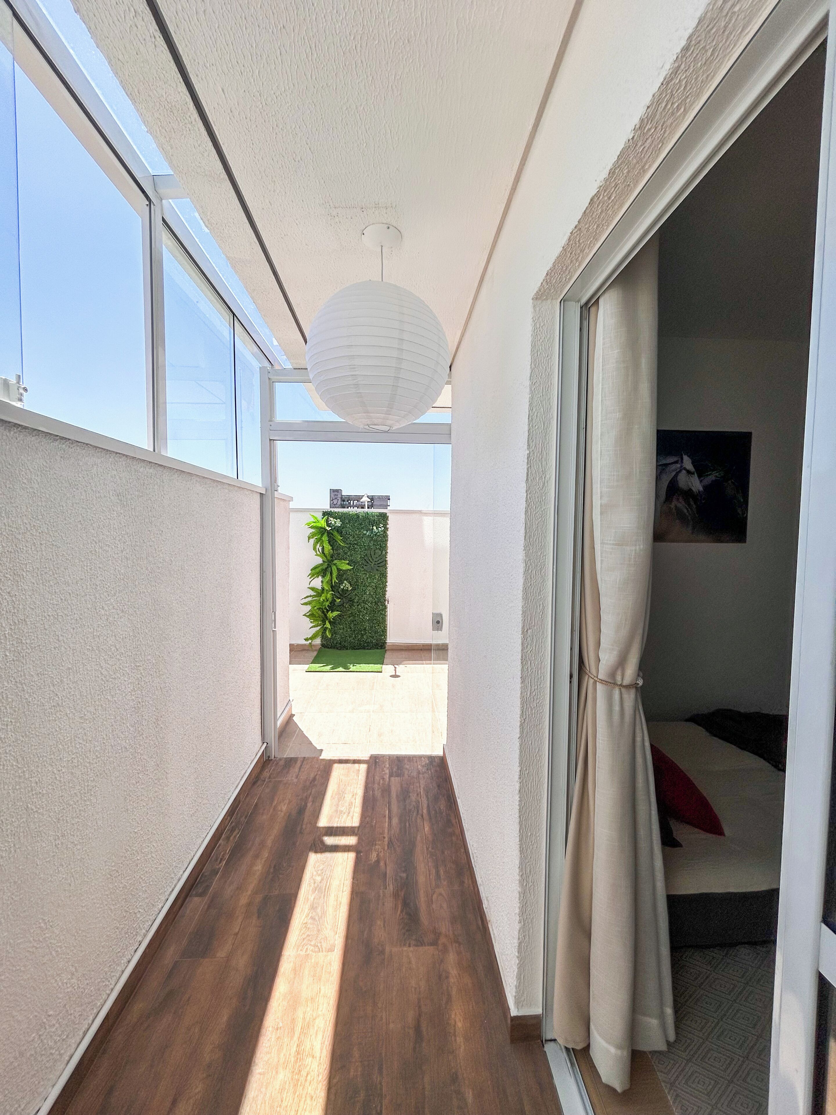 Un pasillo estrecho con suelo de madera, techo blanco y vistas a una pared verde y plantas afuera.