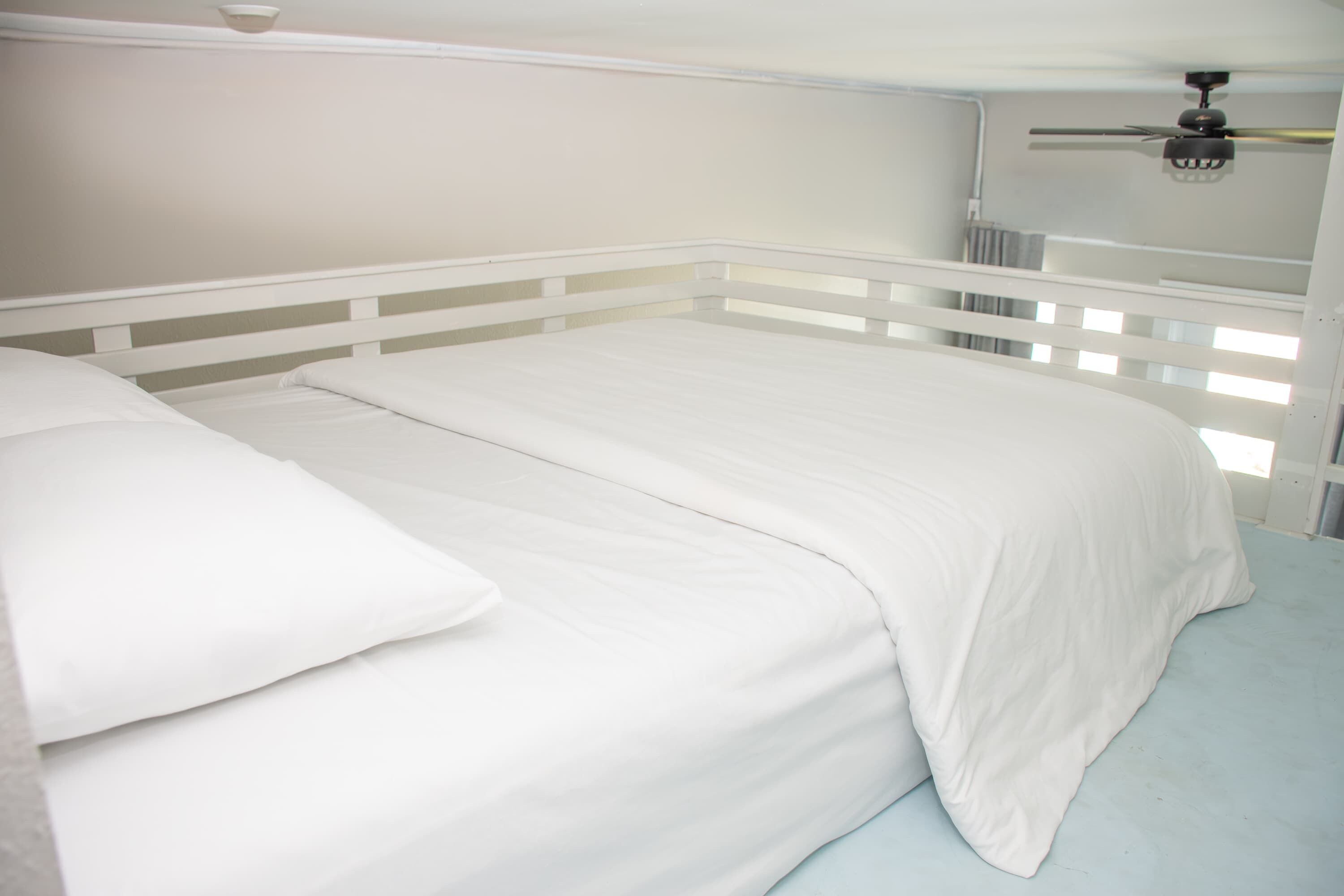 Una cama con ropa de cama blanca en una habitación con paredes y ventilador de techo blancos.