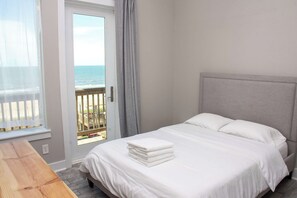 A bedroom with a bed, a wooden table, a balcony with a view of the sea, and a door with curtains.