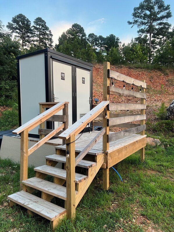 Un baño portátil exterior con una rampa de madera que lo lleva a él, rodeado de césped y árboles.
