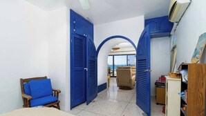Une pièce comprenant un lit, une chaise, un bureau et donnant sur un balcon par une porte ouverte.