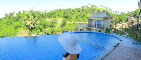 Uma pessoa está de pé perto de uma piscina, com vista para uma encosta e um edifício ao fundo.