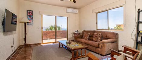 A living room with a brown sofa, wooden coffee table, and chairs, a ceiling fan, and a large sliding glass door leading to a balcony.