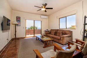 Ein Wohnzimmer mit braunem Sofa, Couchtisch aus Holz und Sesseln, einem Deckenventilator und einer großen Schiebetür, die auf einen Balkon führt.
