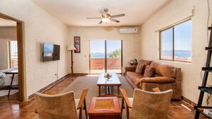 A living room with a sofa, a TV, a ceiling fan, and a view of the sea.
