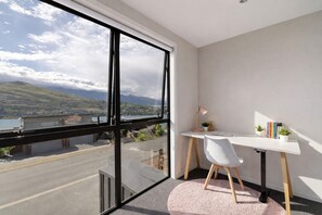 A modern room with a large window, a desk, a chair, and a view of a coastal landscape. - Highview Vista - Lake views, Outdoor Fireplace, great for families (Queenstown)