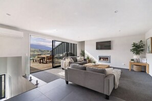 A modern living room with a grey sofa, a fireplace, and a large window overlooking a deck. - Highview Vista - Lake views, Outdoor Fireplace, great for families (Queenstown)