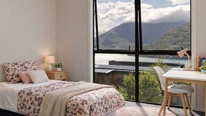 Quarto com uma janela grande, cama com roupa de cama floral, mesa de cabeceira com abajur e escrivaninha com cadeira.