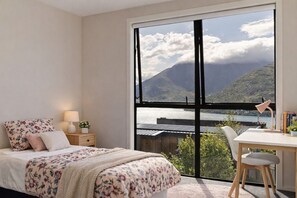 A bedroom with a large window, a bed with floral bedding, a bedside table with a lamp, and a desk with a chair. - Highview Vista - Lake views, Outdoor Fireplace, great for families (Queenstown)