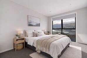 A bedroom with a large bed, two bedside tables, a nightstand, a view of a lake through a large window, and a mountain landscape painting on the wall. - Highview Vista - Lake views, Outdoor Fireplace, great for families (Queenstown)