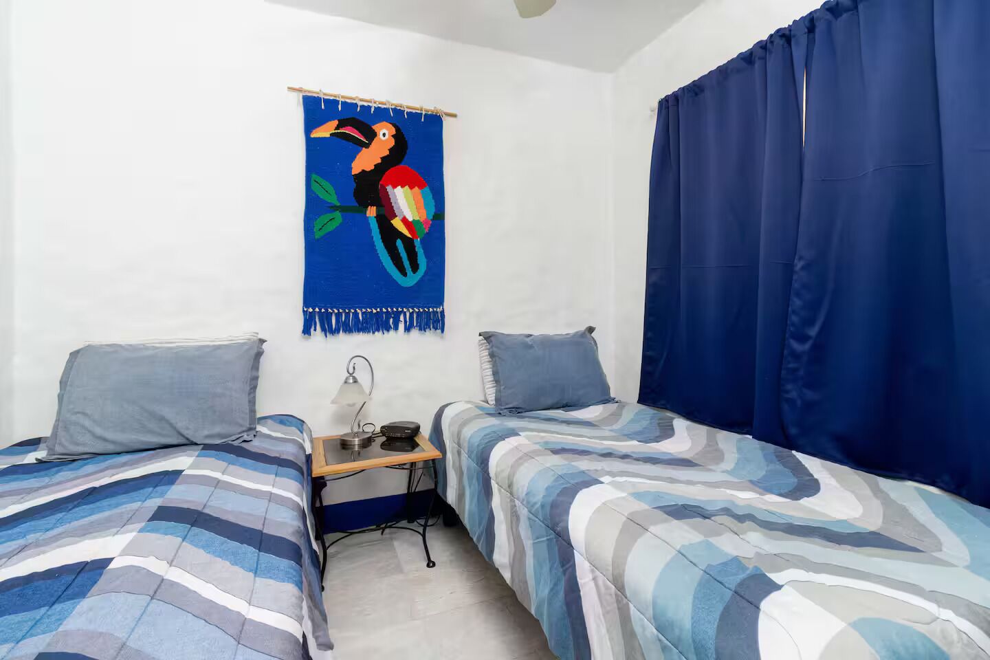 Une petite pièce simple, avec deux lits, un bureau doté d’une lampe et une tapisserie colorée accrochée au mur.