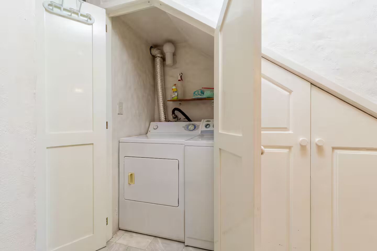 Une buanderie équipée d’un lave-linge et d’un sèche-linge, d’une conduite de ventilation et d’une étagère contenant des produits de nettoyage.