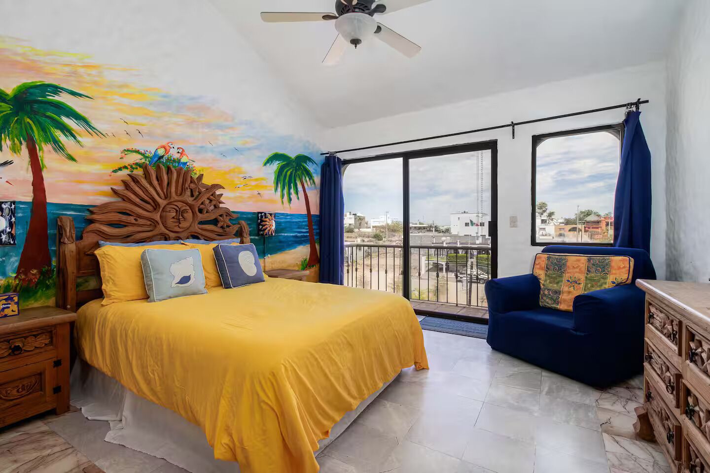A bedroom with a bed, a chair, a nightstand, a wooden headboard, a ceiling fan, and a large window with curtains.