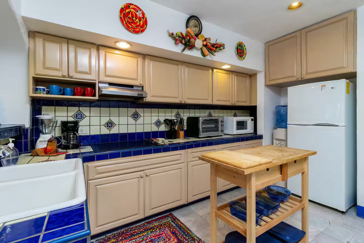Une cuisine avec des meubles de couleur beige, un plan de travail bleu recouvert de carreaux, une table en bois et un réfrigérateur.