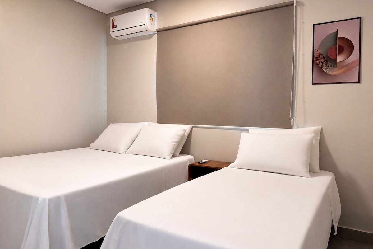 A hotel room with two single beds, a wall-mounted air conditioner, and a framed abstract artwork.