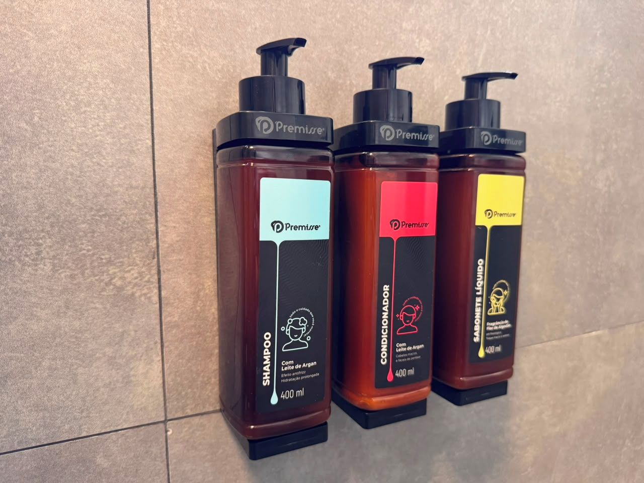 Tres botellas de champú con dosificadores, cada una de un color diferente, frente a una pared de azulejos.