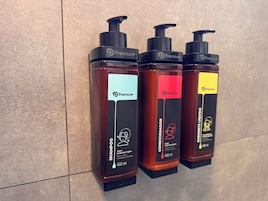 Três frascos de shampoo com bombas dosadoras, cada um em cores diferentes, diante de uma parede revestida com azulejos.