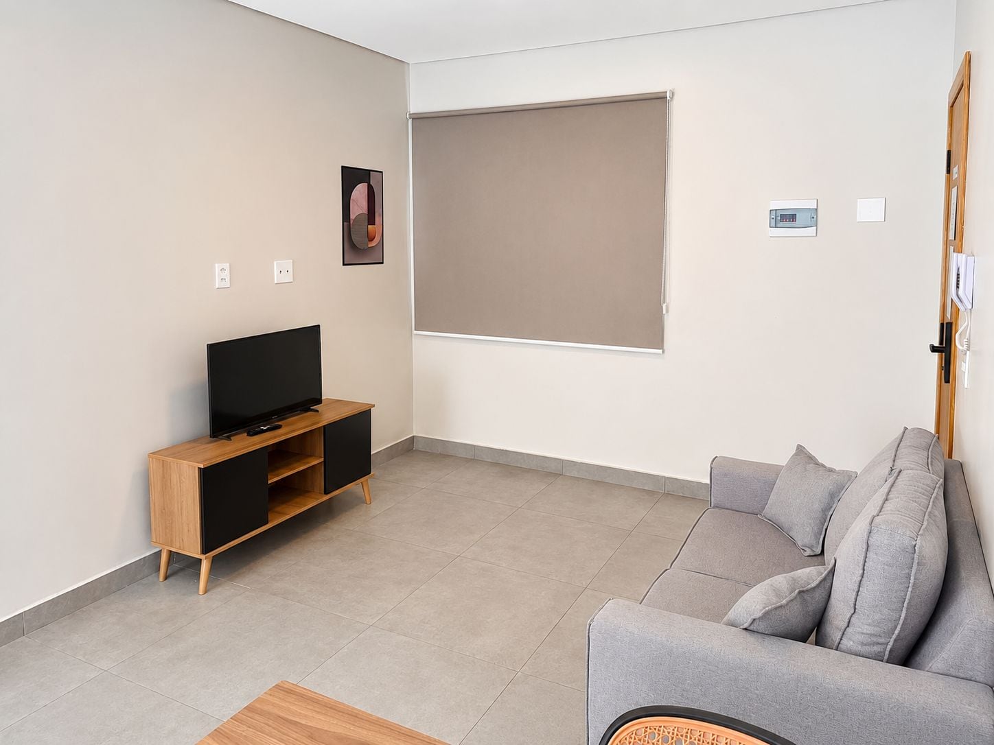 Una sala de estar moderna con un televisor de pantalla plana sobre un soporte de madera, un sofá gris y un ventanal con persiana beige.